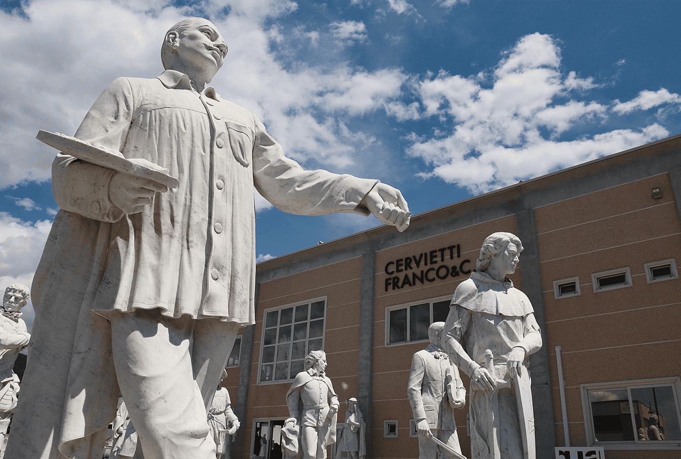 Several white marble statues of people stand outside a building labeled Cervietti Franco & C., under a partly cloudy sky. One statue in the foreground is holding a book or tablet.