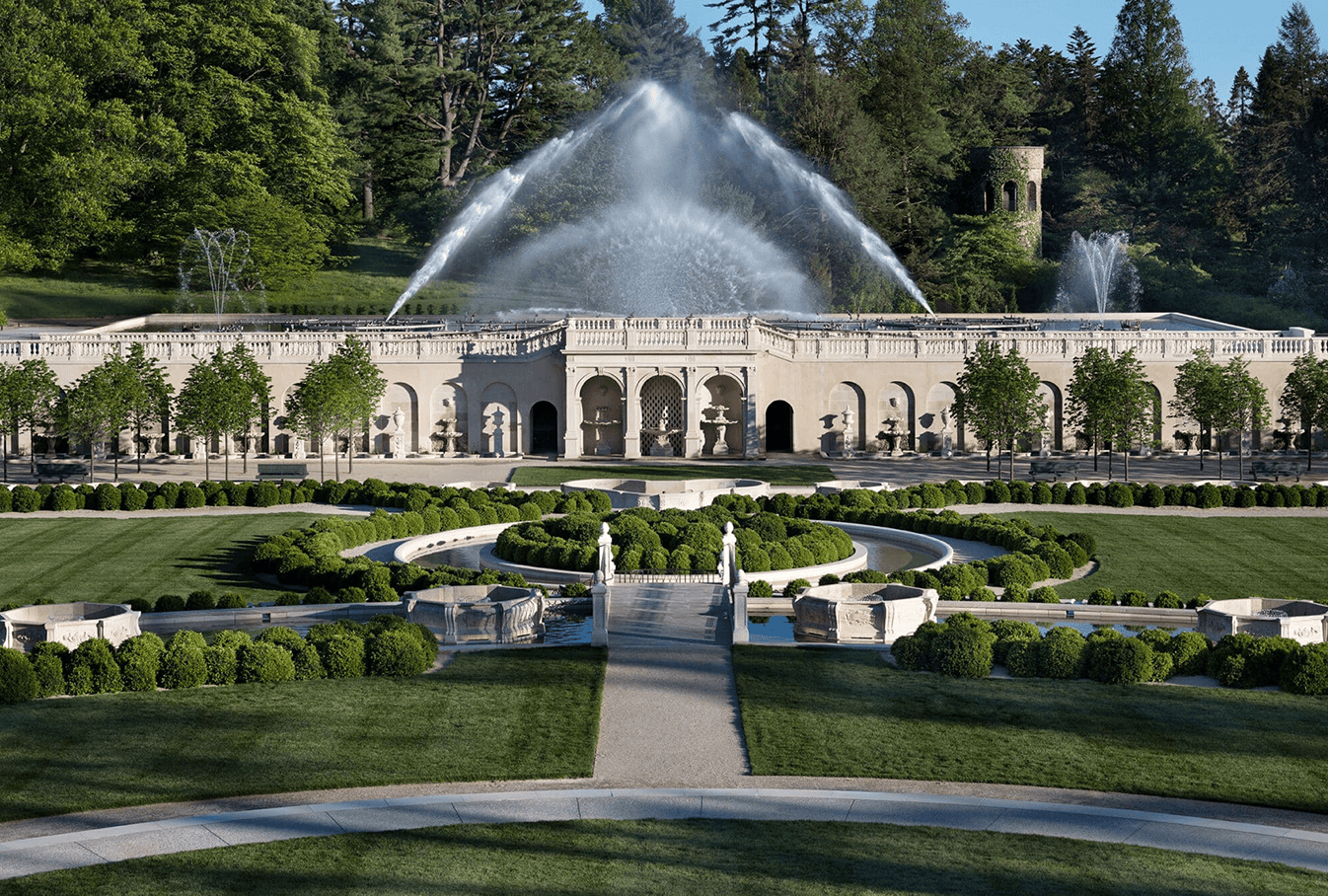 A grand formal garden with symmetrical hedges, a central statue, and a large fountain spraying water high above a stone building surrounded by lush trees.
