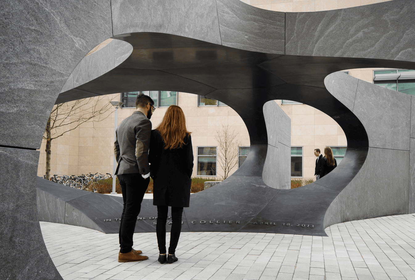 Two people stand in front of a modern, abstract stone and metal sculpture, while another person walks in the background near a building with large windows.