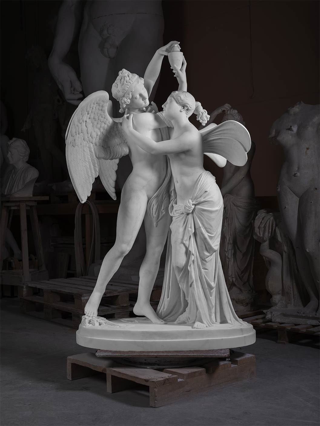 A marble statue of two figures, one winged, holding hands and gazing at each other, displayed in a dimly lit room with other sculptures in the background.