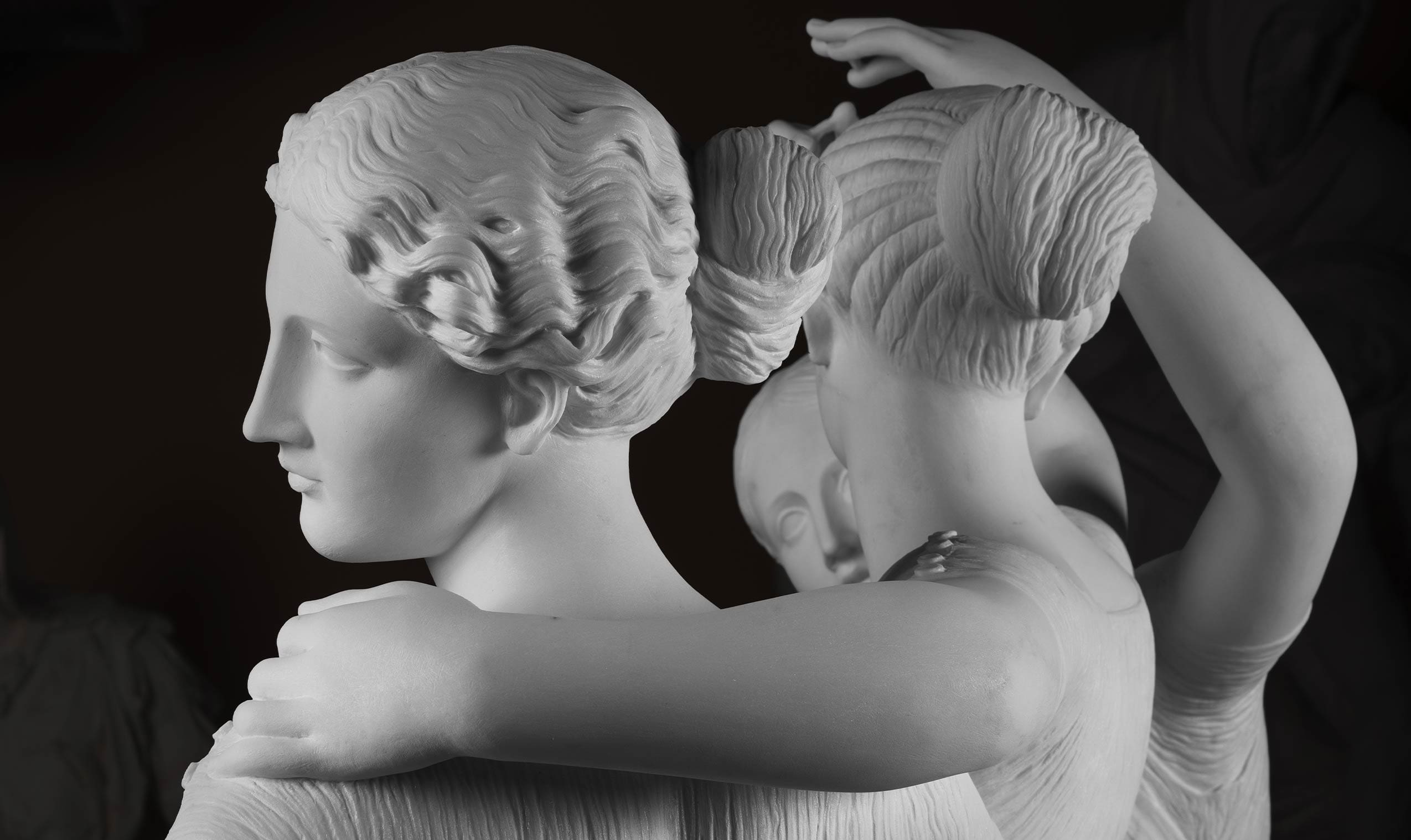 Close-up of a white marble sculpture showing three women with styled hair, arms gently draped over each others shoulders, against a dark background.