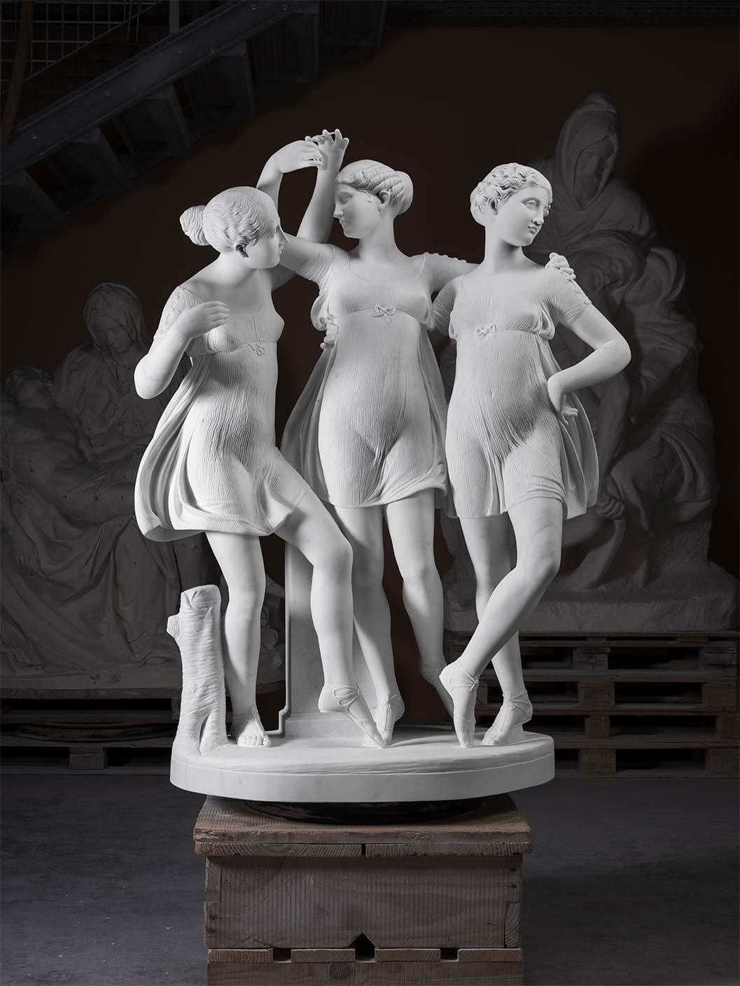 A marble sculpture of three women in sheer dresses, standing closely together and holding hands, displayed on a round base atop wooden pallets in an indoor setting.