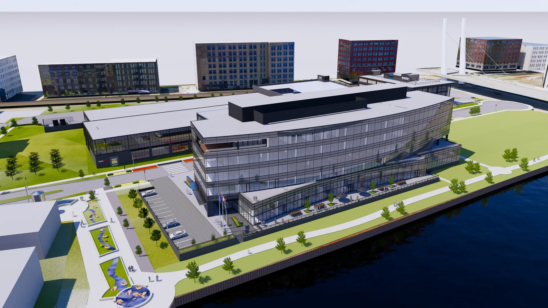 A modern multi-story glass building with parking lot, surrounded by other buildings and green spaces, situated beside a body of water. Pathways and landscaping are visible around the property.