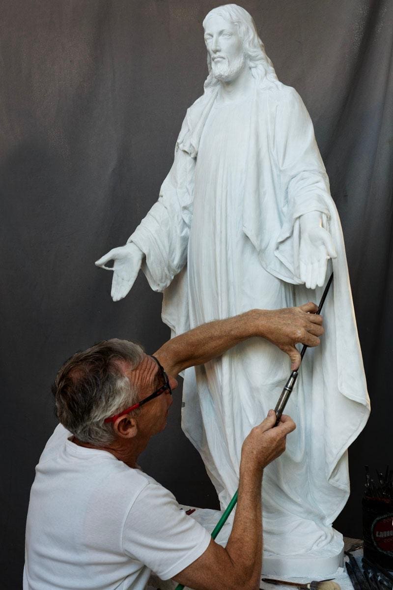 An artist wearing glasses works on a white statue of Jesus, painting details on the robe with a brush. A gray backdrop is behind them.